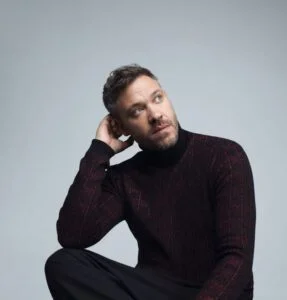 Will Young in a dark jumper, hand to his head