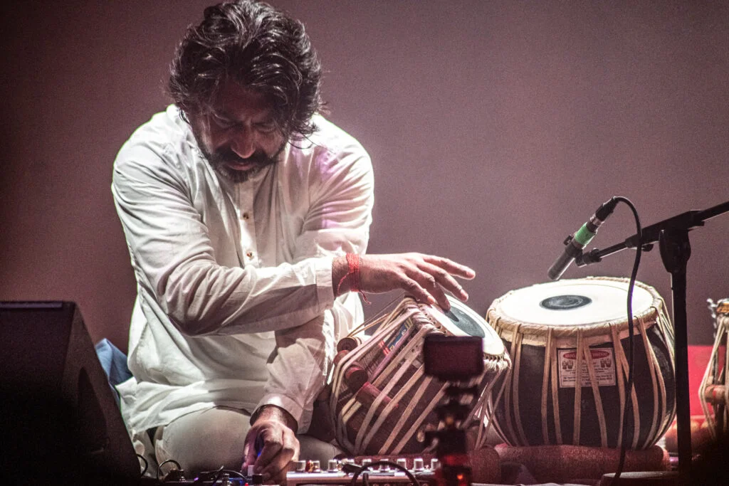 A tabla player in white performing beside a microphone at the Tung Auditorium