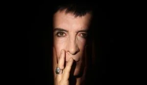Marc Almond with a hand to his face against a black background