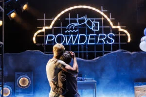 Two actors embracing beneath a neon Powders sign in My Beautiful Laundrette