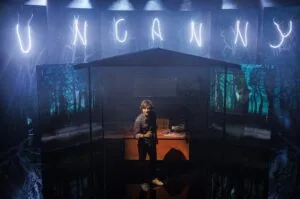 A lone performer on a dark stage beneath the Uncanny sign, photo by Kieran Vyas