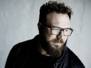 Ben Ottewell in glasses, portrait against a pale wall