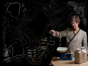 A performer pours from a jar at a table with weighing scales, beside an illustrated map