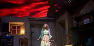 An actor in a long dress on a dark farmhouse set under a red sky in The Croft