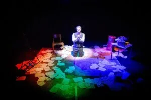 A lone actor kneels on a stage strewn with papers in rainbow light at Hope Street Theatre
