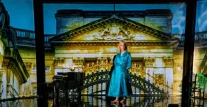 Kit Green as Mrs Dalloway in a blue coat before a projected grand building at Storyhouse